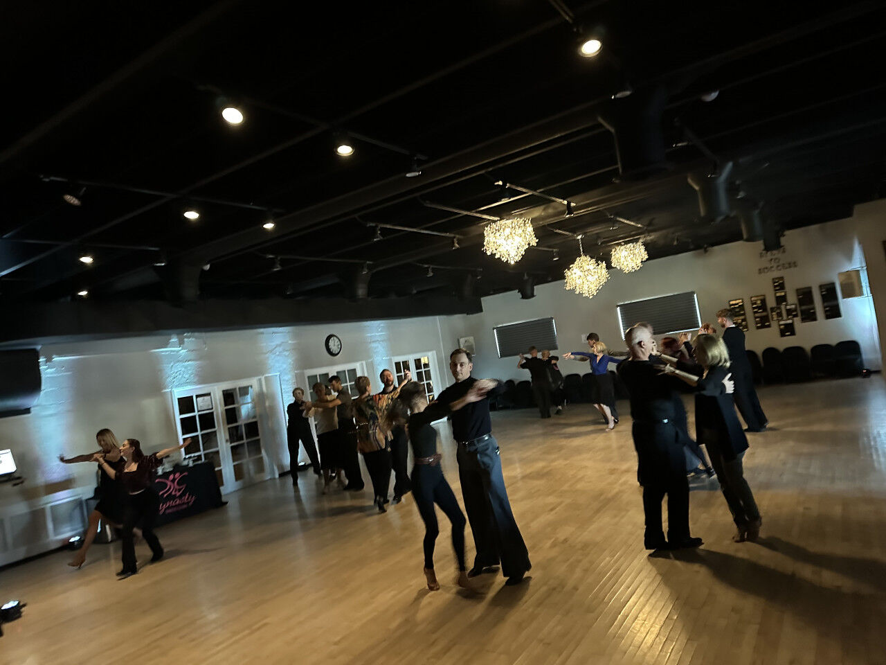 Couples dancing across the wooden floor at a practice party, chandeliers overhead